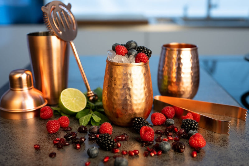 Fruchtiger Beeren-Cocktail in einem stilvollen Kupferbecher, dekoriert mit frischen Beeren, Minze und Limettenscheiben. Im Hintergrund Cocktail-Accessoires wie ein Shaker und frische Zutaten für den perfekten Sommerdrink.